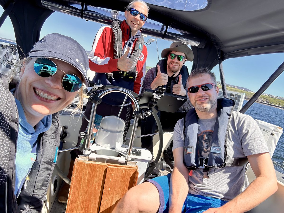 Crew selfie - four sailors in the cockpit, life vests on, thumbs up